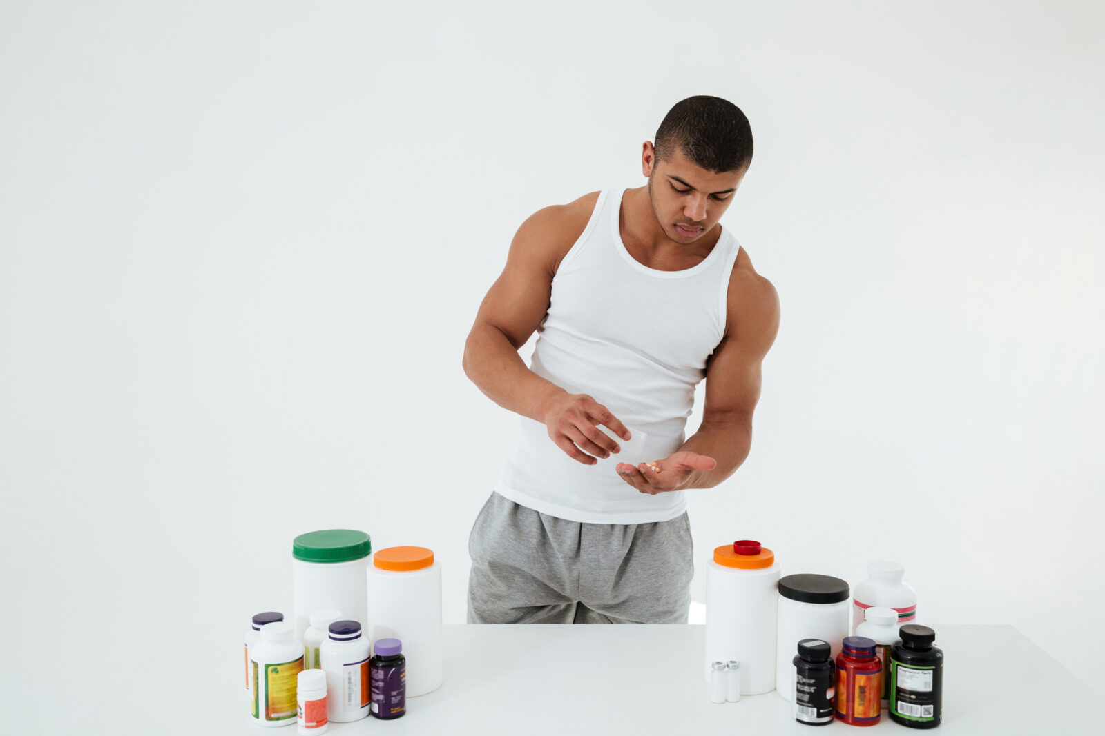 Picture of young sportsman standing over white background holding vitamins and sport pills. Looking aside.